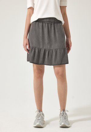 Kvinde iført grå nederdel med elastisk talje og flæsekant, hvid kortærmet top og sølvfarvede sneakers, stående mod en ensfarvet baggrund.