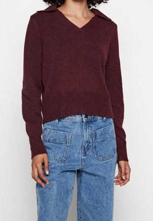 Frau trägt einen burgunderroten, langärmeligen Pullover mit V-Ausschnitt und Kragen sowie hoch taillierte hellblaue Jeans mit aufgesetzten Fronttaschen.