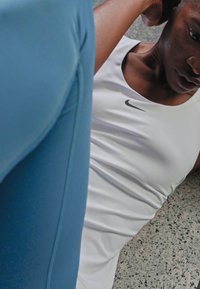 Nike sportska odjeća u bijeloj boji, s bezrukavim dizajnom i suptilnim logotipom. Pozadina prikazuje strukturirani pod i plavi odjevni predmet.