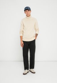 Beige polo sweater med ribbede manchetter og nederste kant, parret med sorte bukser og lyse sneakers. En blå denimkasket fuldender outfittet.