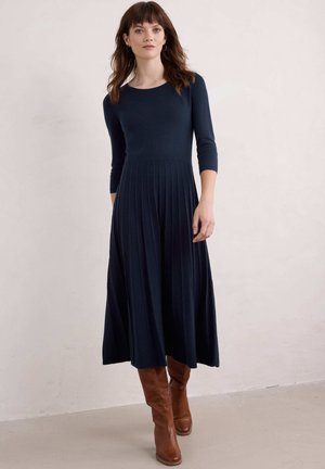 Navyblaues, gestricktes Midi-Kleid mit einem runden Halsausschnitt, 3/4-Ärmeln und plissiertem Rock. Tragen mit braunen Lederstiefeln bis zum Knie.