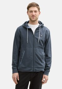 Marineblå hættetrøje med lynlås i en tekstureret stof, to forlommer og kontrastfarvede snore i hætten. Båret sammen med en hvid t-shirt.