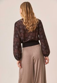Blusa de manga larga en tela oscura con patrones intrincados, combinada con pantalones anchos de pierna recta, ligeros y brillantes. Presenta mangas fruncidas y una cinturilla ajustada.