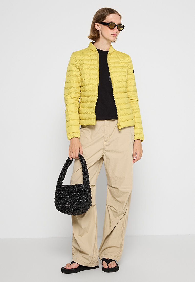 Gelbe Puffjacke, leicht, mit Reißverschluss. Schwarzes T-Shirt, beige weite Hose. Schwarze strukturierte Handtasche, flache Sandalen, übergroße Sonnenbrillen.