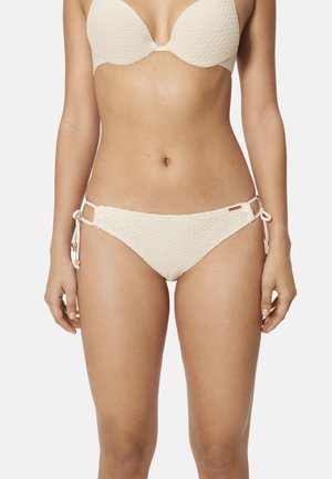 Mujer con bikini texturizado color blanco roto con braguitas con lazo lateral y top con aros y relleno, sobre fondo liso.