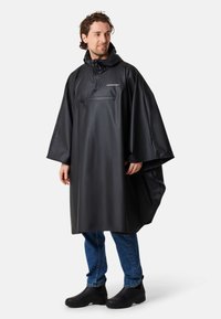 Svart vattentät poncho med huva, med en frampocket med dragkedja och vida ärmar, bärs över jeans och svarta gummistövlar.
