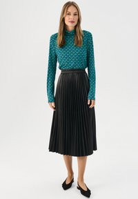 Koszula z długim rękawem w odcieniu teal z zielonym i niebieskim wzorem, zestawiona z czarną spódnicą plisowaną i czarnymi butami typu slingback.