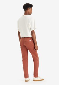 Pantalons couleur rouille avec une coupe slim, dotés de deux poches arrière et d'une étiquette discrète. Associés à une chemise blanche à manches courtes ample.