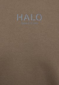 Brunt bomullstyg med en ribbad textur och en ljusblå "HALO"-logotyp samt åren "1968 1972 1981" tryckta nedanför.