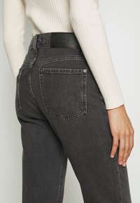 Primer plano de una persona que lleva jeans oscuros de talle alto y un top de manga larga acanalado en color crema, con la mano reposando a un lado.