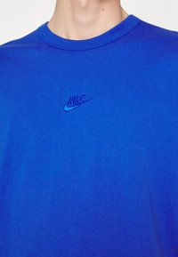 Modré bavlněné tričko s kulatým výstřihem. Na hrudi má vyšívané logo Nike v mírně tmavší modré barvě. Jemná textura.