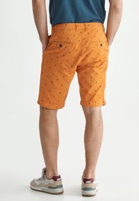 Shorts orange en tissu léger, avec de petits motifs sombres, deux poches arrière avec des boutons, et une coupe droite. Associés à des baskets.