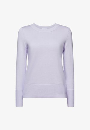 Femme aux longues tresses portant un pull lavande, tenant un sac à main bordeaux à motif croco et une veste vert foncé nouée à la taille.
