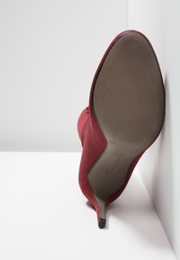 Bottine chaussette rouge à talon haut reposant la semelle vers le haut contre un mur et un sol blancs, montrant la semelle en caoutchouc texturée et le bout pointu.