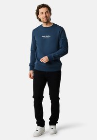 Felpa blu navy con collo a giro, logo testuale sul davanti, polsini e orlo a costine. Abbinata a pantaloni neri e scarpe bianche.