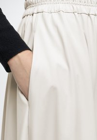 Main dans la poche d'une jupe taille élastique beige clair portée avec un haut noir à manches longues.