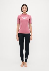 Rash guard rosa a maniche corte con logo bianco sopra leggings neri, caratterizzato da una texture liscia e una vestibilità atletica.