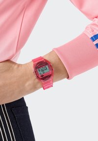 Skaitmeninis laikrodis su aiškiu rožiniu plastiko korpusu ir atitinkančia juosta, turintis juodą skystųjų kristalų ekraną ir baltus žymėjimus. Nešiojamas ant riešo.