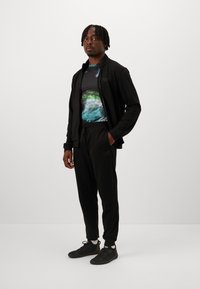 Schwarze Zip-Jacke und Hose, kombiniert mit einem mehrfarbigen Grafik-T-Shirt mit blauen und grünen Mustern, getragen mit schwarzen Sportschuhen.