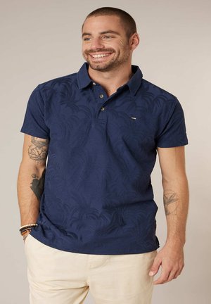 Glunderende man met kort haar, gekleed in een marineblauw poloshirt met structuur en beige broek, met tatoeages op beide onderarmen en kralenarmbanden.