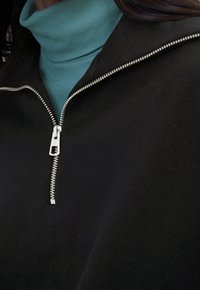 Czarna kurtka na zamek błyskawiczny z srebrnymi detalami, noszona na turkusowym golfie. Gładka tekstura, nowoczesny design i dopasowany krój.