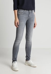 Kapeeden farkut vaaleassa harmaassa denimissä, jossa on sileä pinta. Varustettu viidellä taskulla, vyölenkeillä ja metallinappikiinnityksellä. Yhdistettynä valkoisiin tennareihin.