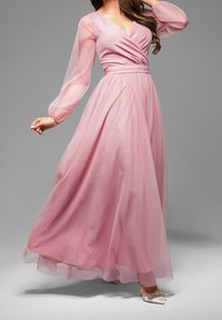 Femme portant une longue robe rose fluide avec des manches longues transparentes et un décolleté en V, posant avec un bras levé et une main tendue.