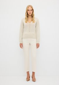 Beige gebreide trui met een V-hals en centrale knoopsluiting, gecombineerd met lichtbeige slim-fit broeken en naakte hoge hakken.