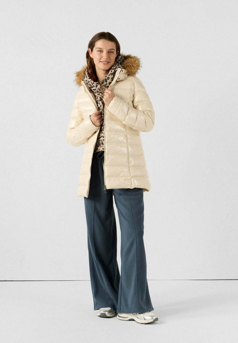 Chaqueta de plumas en crema con capucha forrada de piel, diseño acolchado y cierre frontal con cremallera. Combinada con pantalones amplios de color azul oscuro y zapatillas.