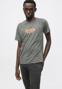 Gray cotton t-shirt featuring an orange "Stranger Things" graphic on the front. Short sleeves and crew neckline, made from soft fabric.