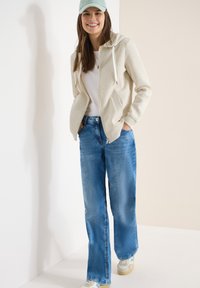 Femme souriante portant un sweat à capuche zippé beige clair, un t-shirt blanc, un jean bleu à jambes larges, des baskets et une casquette vert pastel, les mains dans les poches.