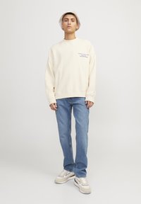 Sweatshirt crème avec texte bleu, coupe loose, accompagné d'un jean bleu clair. Pour parfaire le look, on y ajoute des baskets neutres et un chapeau seau beige.