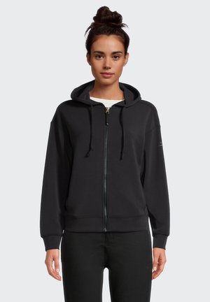 Frau mit dunkel gebundenen Haaren, die eine schwarze Kapuzenjacke mit Reißverschluss über einem weißen Hemd und schwarze Hose trägt, schaut frontal vor einem schlichten Hintergrund.