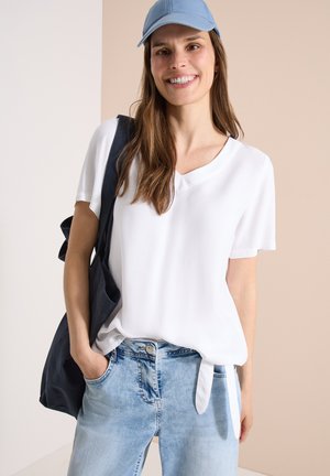 Joven sonriente, lleva una gorra azul clara, una camiseta blanca de manga corta con lazada delante, vaqueros azul claro y lleva un bolso negro de hombro.