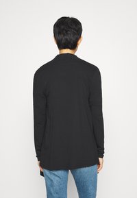 Persona con capelli neri corti che indossa una maglietta nera a maniche lunghe e jeans blu, vista da dietro su uno sfondo bianco semplice.