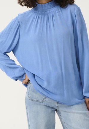 Kobieta ubrana w jasnoniebieską, fakturowaną bluzkę z długim rękawem i marszczonym wysokim kołnierzem oraz jasne dżinsy typu denim, ręka oparta na biodrze.