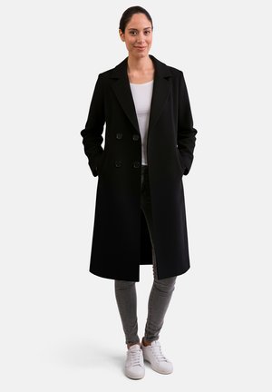 Cappotto nero doppiopetto con grandi bottoni, maniche lunghe e tasche laterali, indossato sopra una maglietta bianca e jeans grigi, abbinato a sneakers bianche.