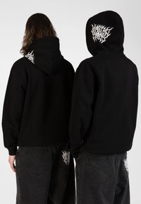 Zwarte capuchonsweaters hebben een grafisch ontwerp op de achterkant en schouders, gecombineerd met donkere jeans die vergelijkbare witte accenten tonen.
