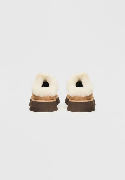 Καφέ σουέτ παπούτσια slip-on με κρέμα γούνα στο εσωτερικό, επίπεδες καφέ καουτσούκ σόλες και στρογγυλή πλάτη, θέα από πίσω.
