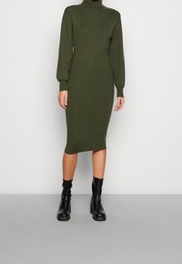 Robe en tricot vert olive avec col montant, manches bouffantes longues et silhouette ajustée, associée à des bottines noires.