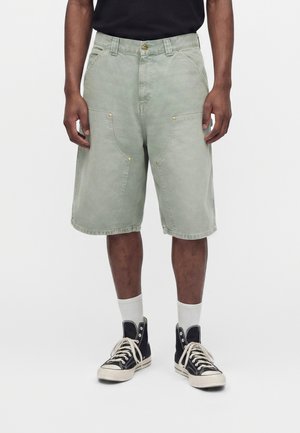 Homme portant un short vert clair jusqu'aux genoux, des baskets montantes noires avec des lacets blancs, des chaussettes blanches et un t-shirt noir raccourci au-dessus de la taille.