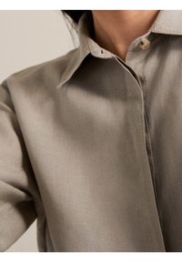 Chemise beige clair structurée en tissu lisse. Elle présente un col pointu et une fermeture à un bouton, avec un détail de couture visible.