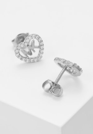 Sølv ørestikker med hjerteformede rammer indlagt med klare sten, med initialerne "MK" i midten, vist på en hvid overflade.