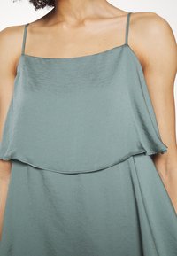 Femme portant une robe sans manches couleur teal clair avec des bretelles fines et un décolleté carré à étages.