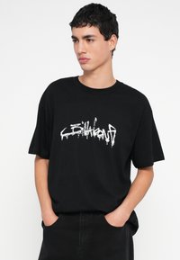 Μαύρο βαμβακερό T-shirt με άνετη εφαρμογή, με λευκό κείμενο σε στυλ γκράφιτι στο μπροστινό μέρος και κοντά μανίκια. Απλό φόντο.