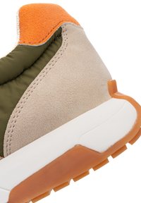 Nahaufnahme einer Sneaker-Ferse mit beigem Wildleder, olivfarbenem Stoff, orangefarbenen Wildlederakzenten und weißer Sohle mit oranger Gummiaußensohle.