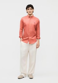 Mężczyzna o kręconych włosach, ubrany w koralową koszulę na guziki, luźne białe spodnie i beżowe mokasyny, stojący na tle jednolitego jasnego tła.