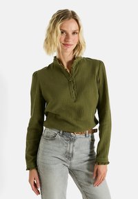 Camisa de manga larga verde oliva con un tejido texturizado, cuello abotonado y puños con volantes, usada con pantalones vaqueros grises claros y un cinturón marrón.