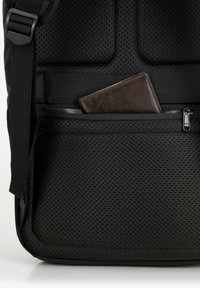 Mochila negra con un bolsillo de malla texturizada. Una billetera de cuero marrón es parcialmente visible en un compartimento con cremallera. Incluye una correa ajustable.