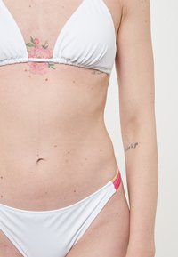 Top bikini triangolare bianco con dettagli arricciati, abbinato a slip bikini bianchi con accenti laterali rosa e marrone chiaro.